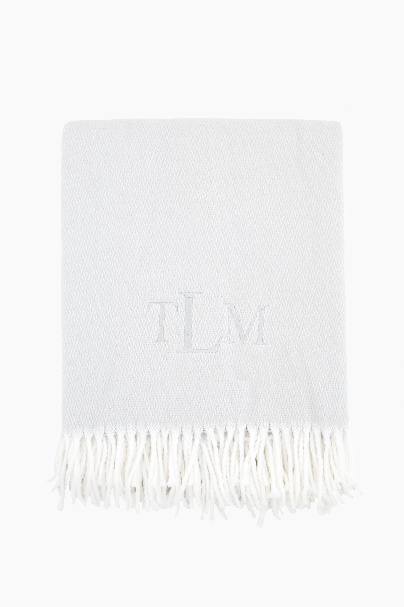 A light grey Matouk Silver Pezzo Throw features a white fringe trim and the monogram TLM embroidered at the center.