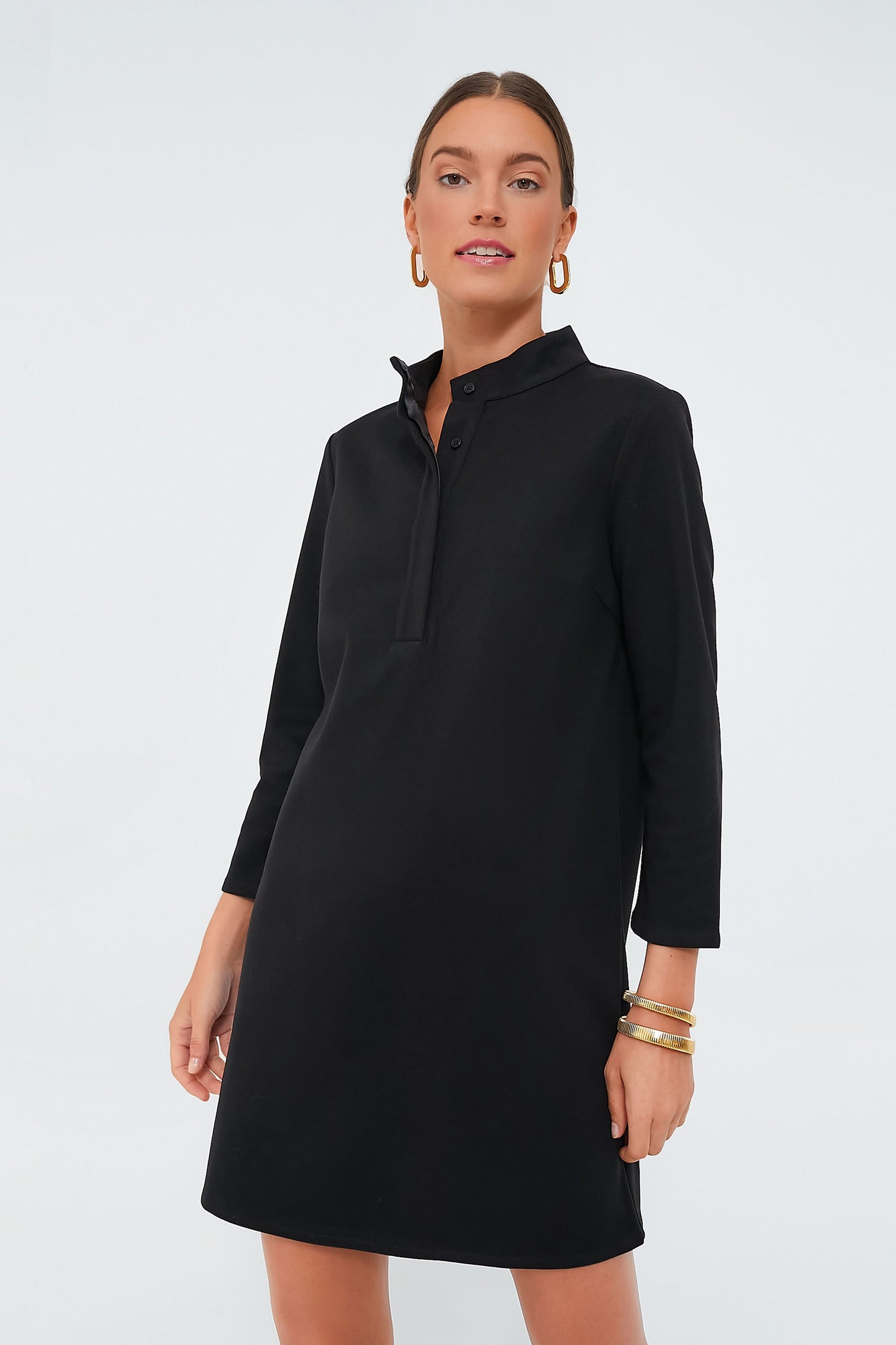 A woman wears a Tuckernuck black long-sleeve stretch knit dress with buttons at the mock neckline, paired with gold hoop earrings and bracelets.