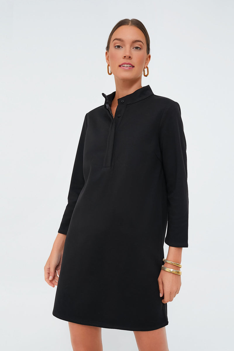 A woman wears a Tuckernuck black long-sleeve stretch knit dress with buttons at the mock neckline, paired with gold hoop earrings and bracelets.