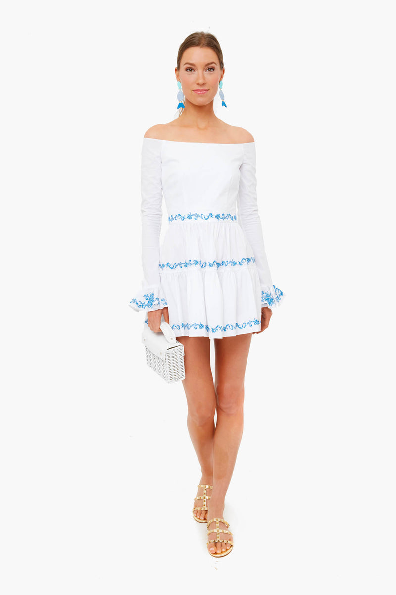A woman faces forward in a Caroline Constas white tiered mini dress with blue embroidery, gold sandals, blue earrings, and a white handbag.