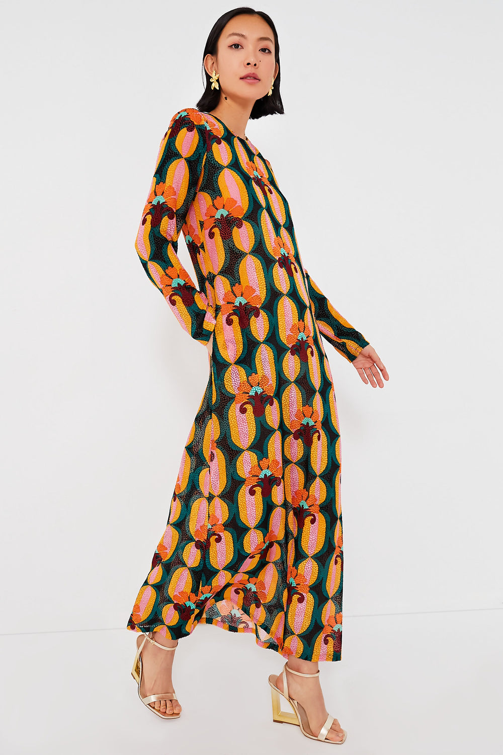 A woman in a bold geometric long sleeve swing dress by La DoubleJ, styled with gold earrings and gold heeled sandals, stands poised.