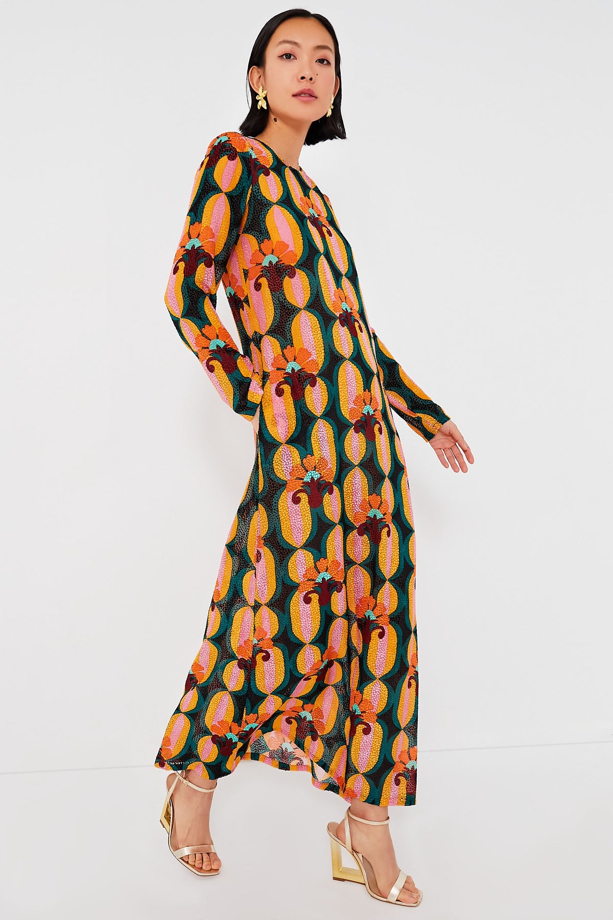A woman in a bold geometric long sleeve swing dress by La DoubleJ, styled with gold earrings and gold heeled sandals, stands poised.