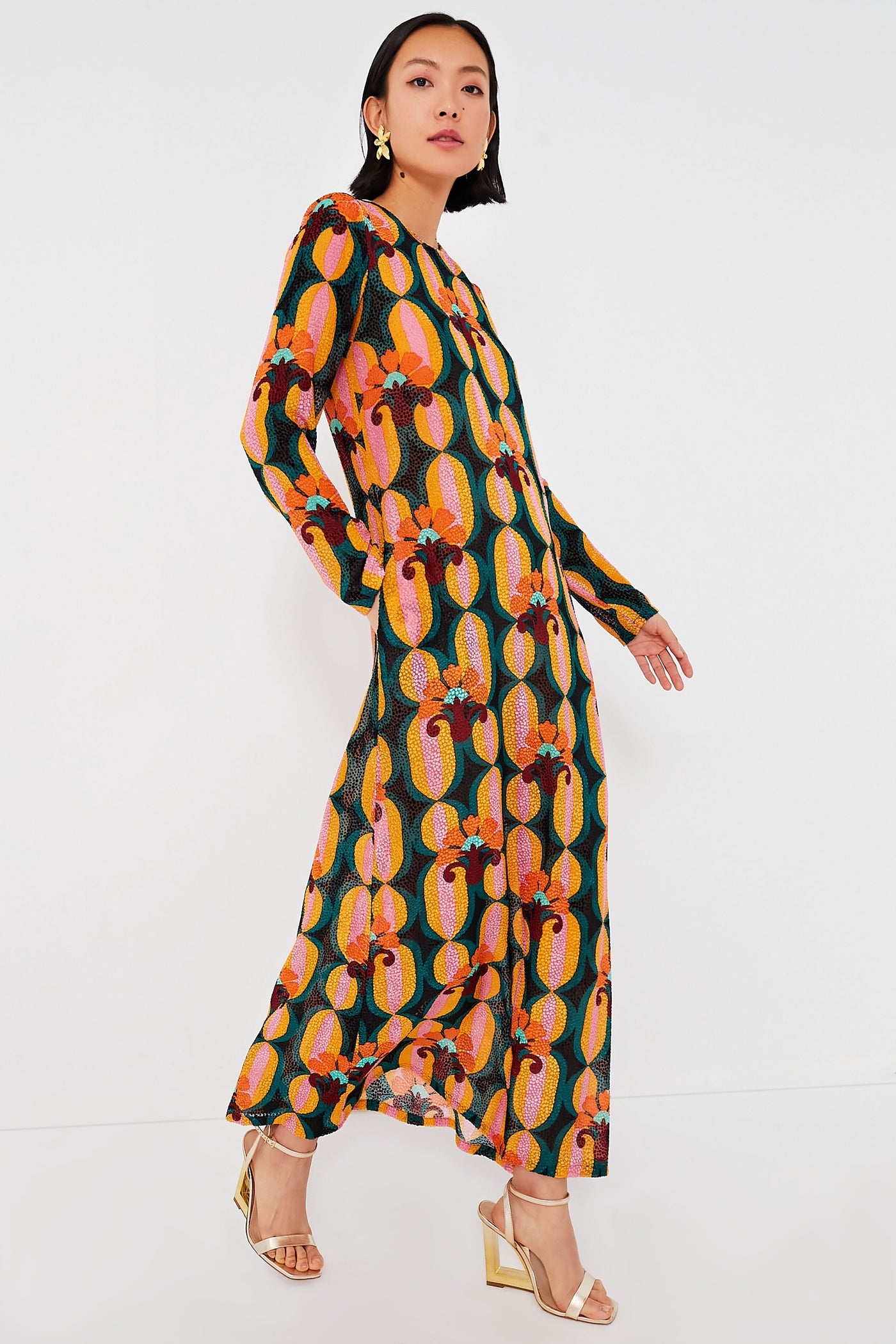 A woman in a bold geometric long sleeve swing dress by La DoubleJ, styled with gold earrings and gold heeled sandals, stands poised.