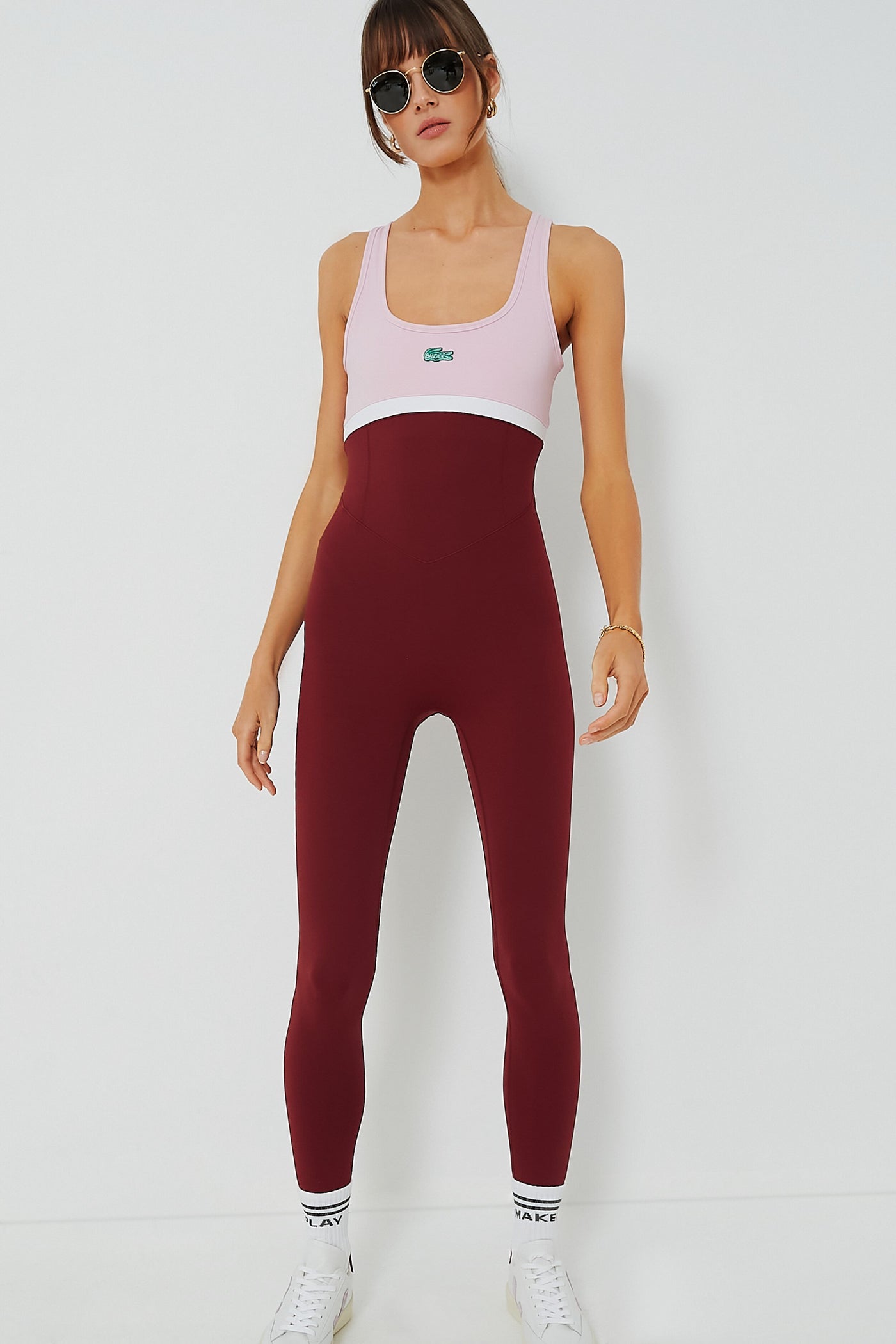 A woman in sunglasses wears a pink and burgundy sleeveless colorblocked one-piece jumpsuit by Bandier x Lacoste, paired with white sneakers and socks, showcasing stylish tennis performance wear.