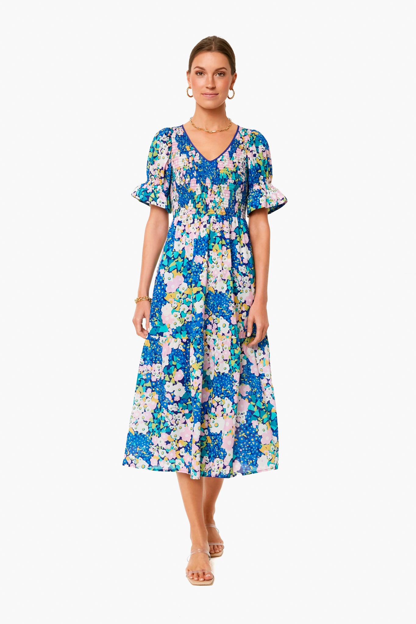 A woman stands wearing a Tuckernuck blue floral print midi dress with a smocked bodice, short ruffled sleeves, gold hoop earrings, a bracelet, and beige flat sandals.