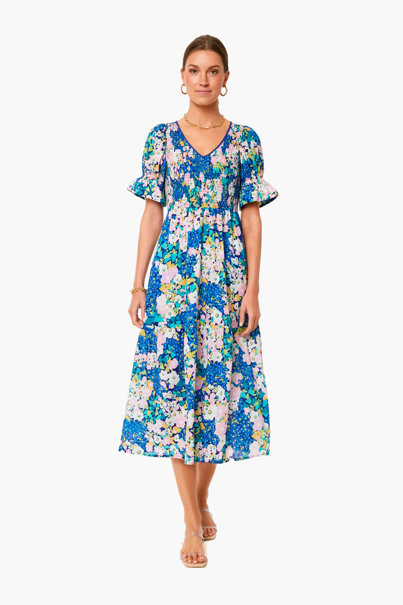 A woman stands wearing a Tuckernuck blue floral print midi dress with a smocked bodice, short ruffled sleeves, gold hoop earrings, a bracelet, and beige flat sandals.