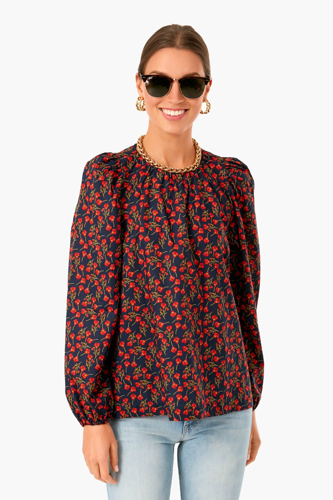 A woman in APOF navy floral print blouse with red and green pattern and balloon sleeves, sunglasses, gold chain necklace, hoop earrings, and light blue jeans.