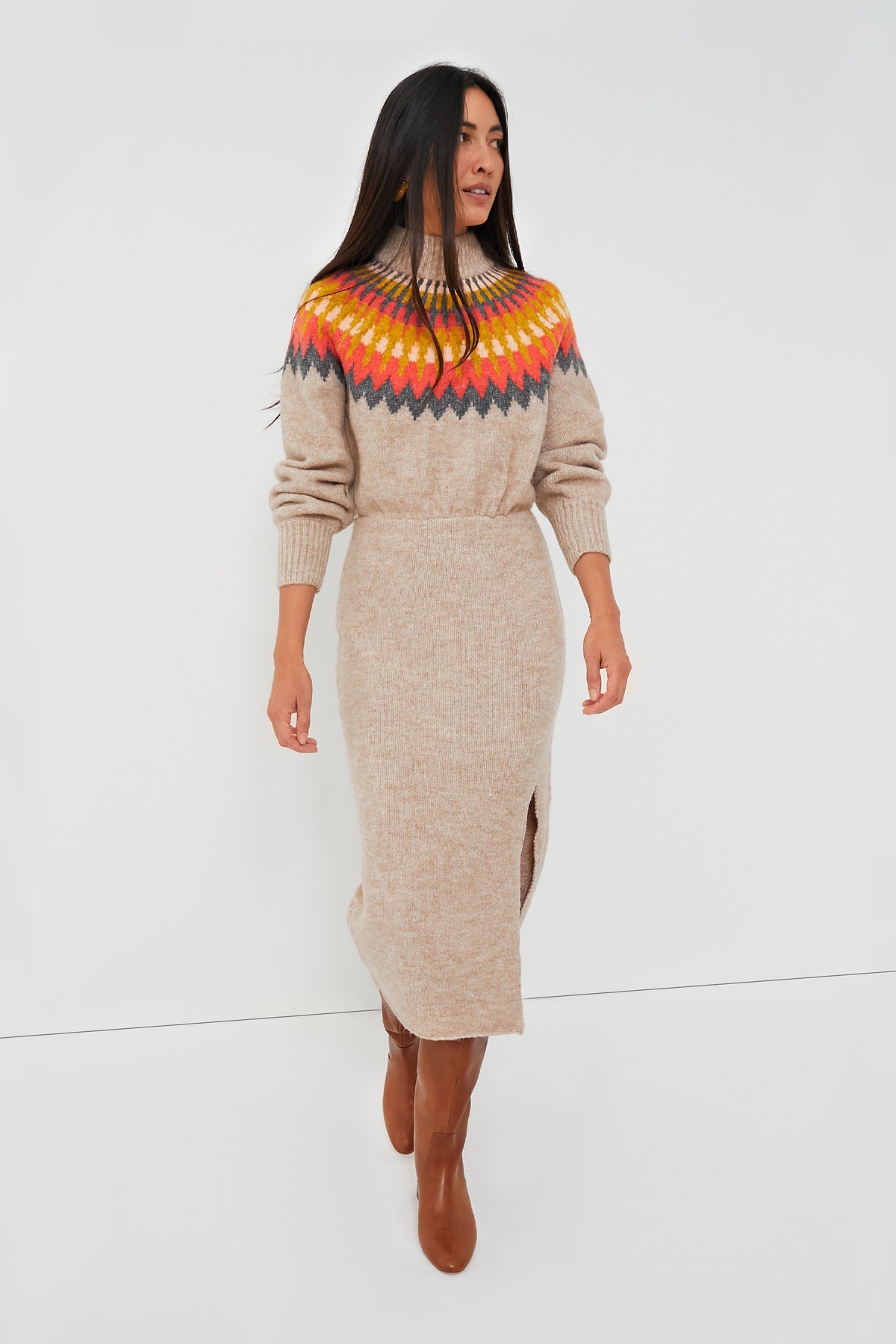 A woman wears a beige Saylor Fair Isle sweater dress featuring a colorful patterned yoke, long sleeves, ribbed turtleneck, and front slit, styled with brown knee-high boots.