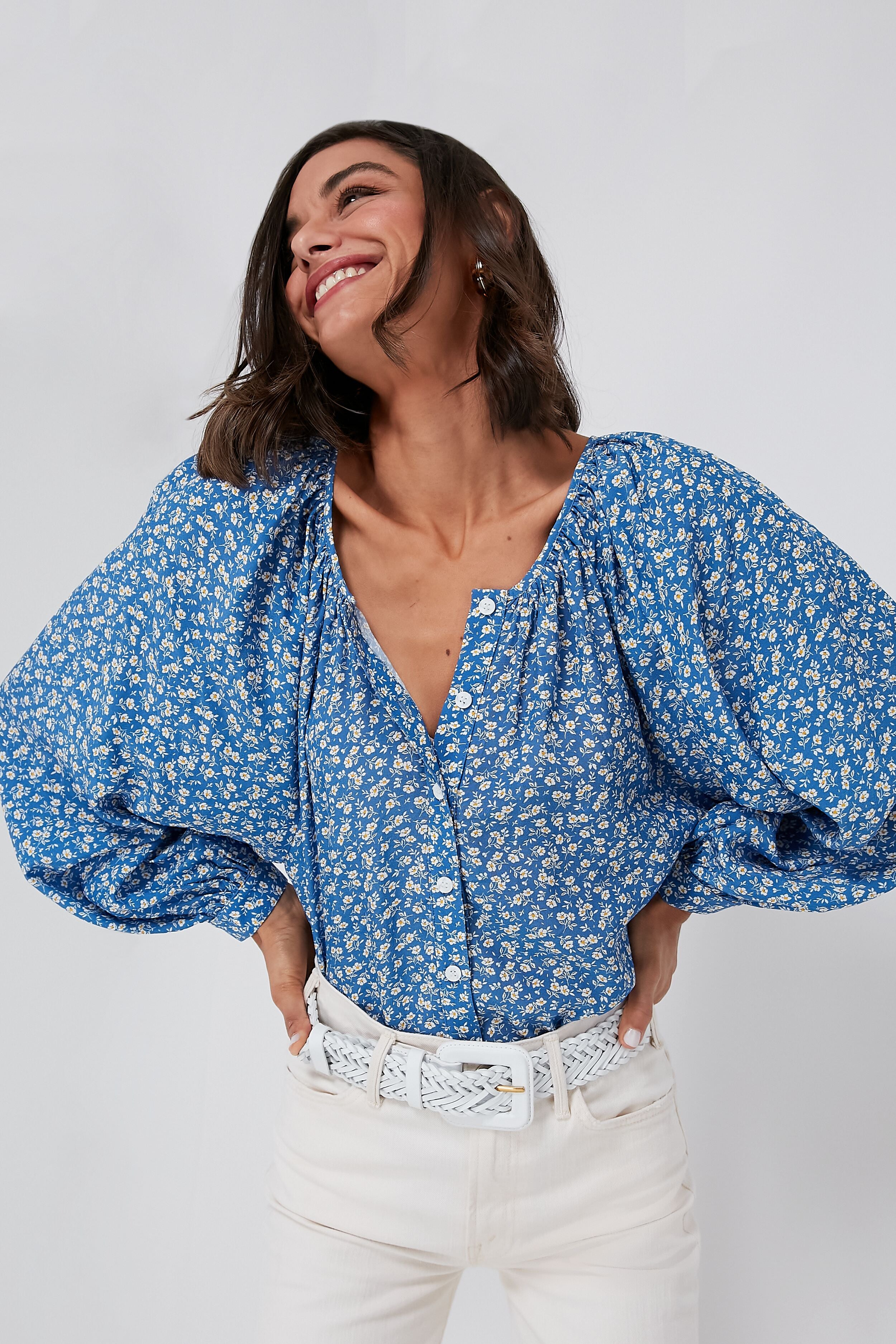 A woman smiles with her hands on her hips, wearing Emerson Fry’s blue boho floral blouse with puffed sleeves and white pants.