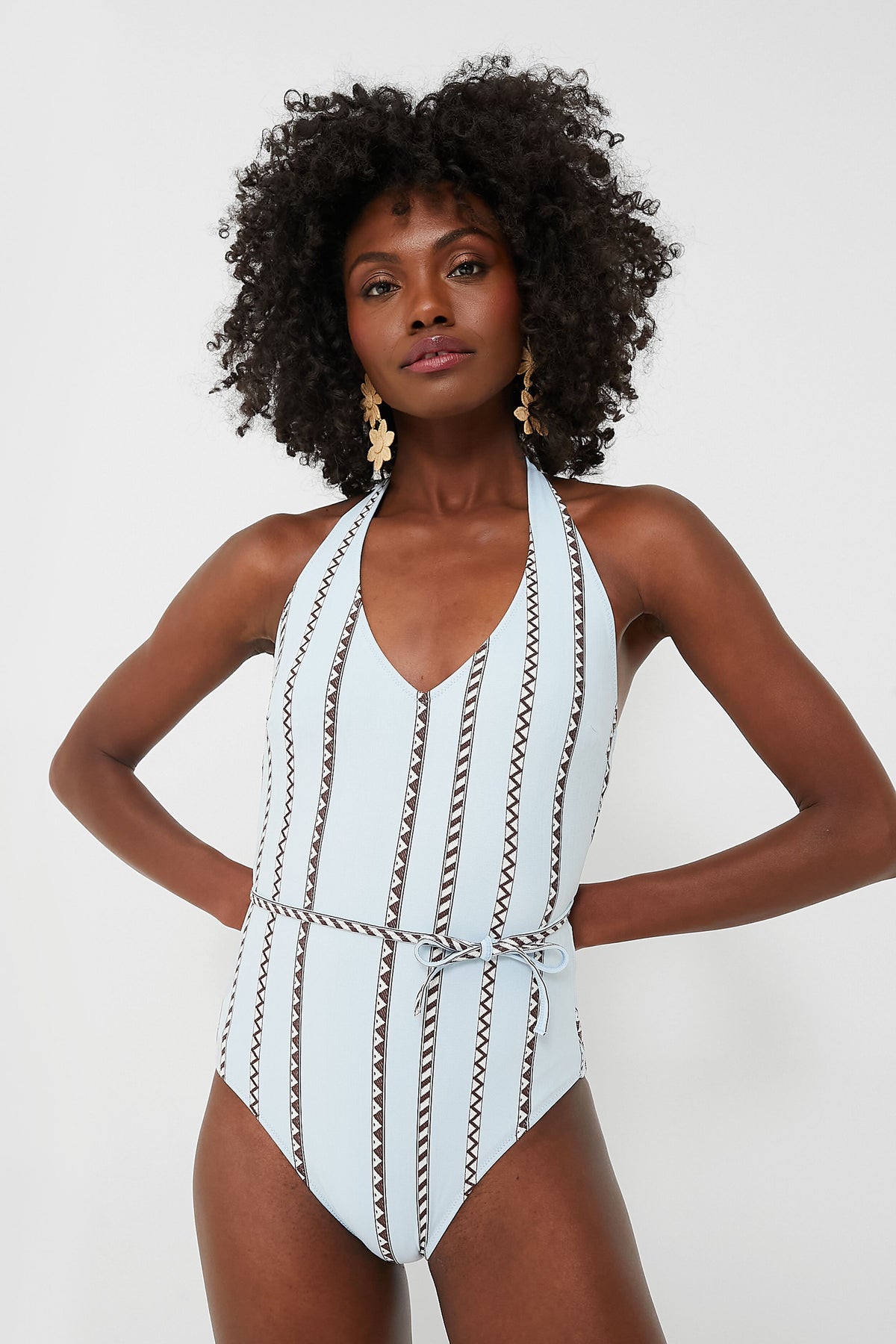 Woman wearing a lemlem light blue patterned one-piece swimsuit with a belted waist, halter neckline, and deep V-neck accent.