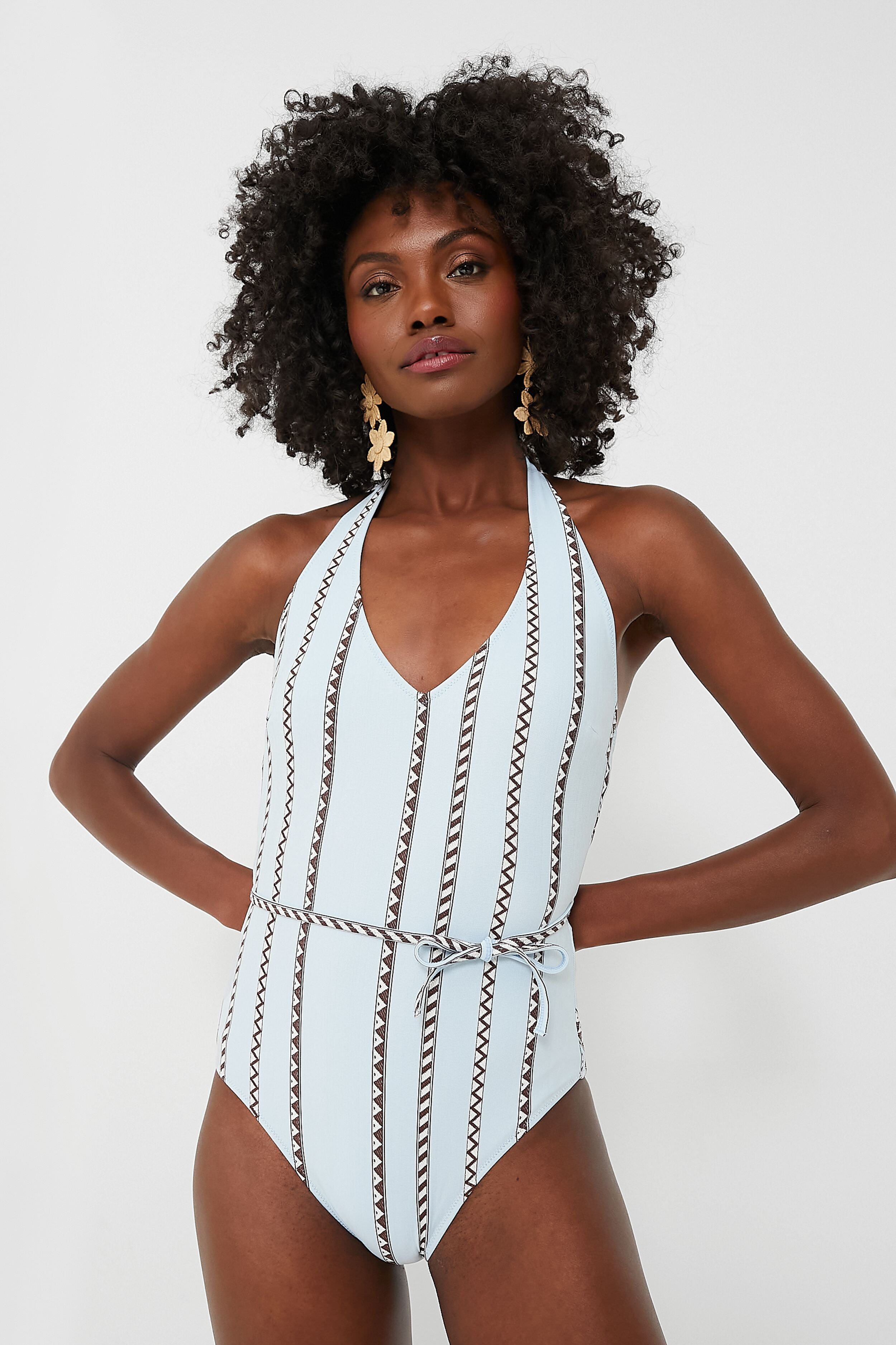 Woman wearing a lemlem light blue patterned one-piece swimsuit with a belted waist, halter neckline, and deep V-neck accent.