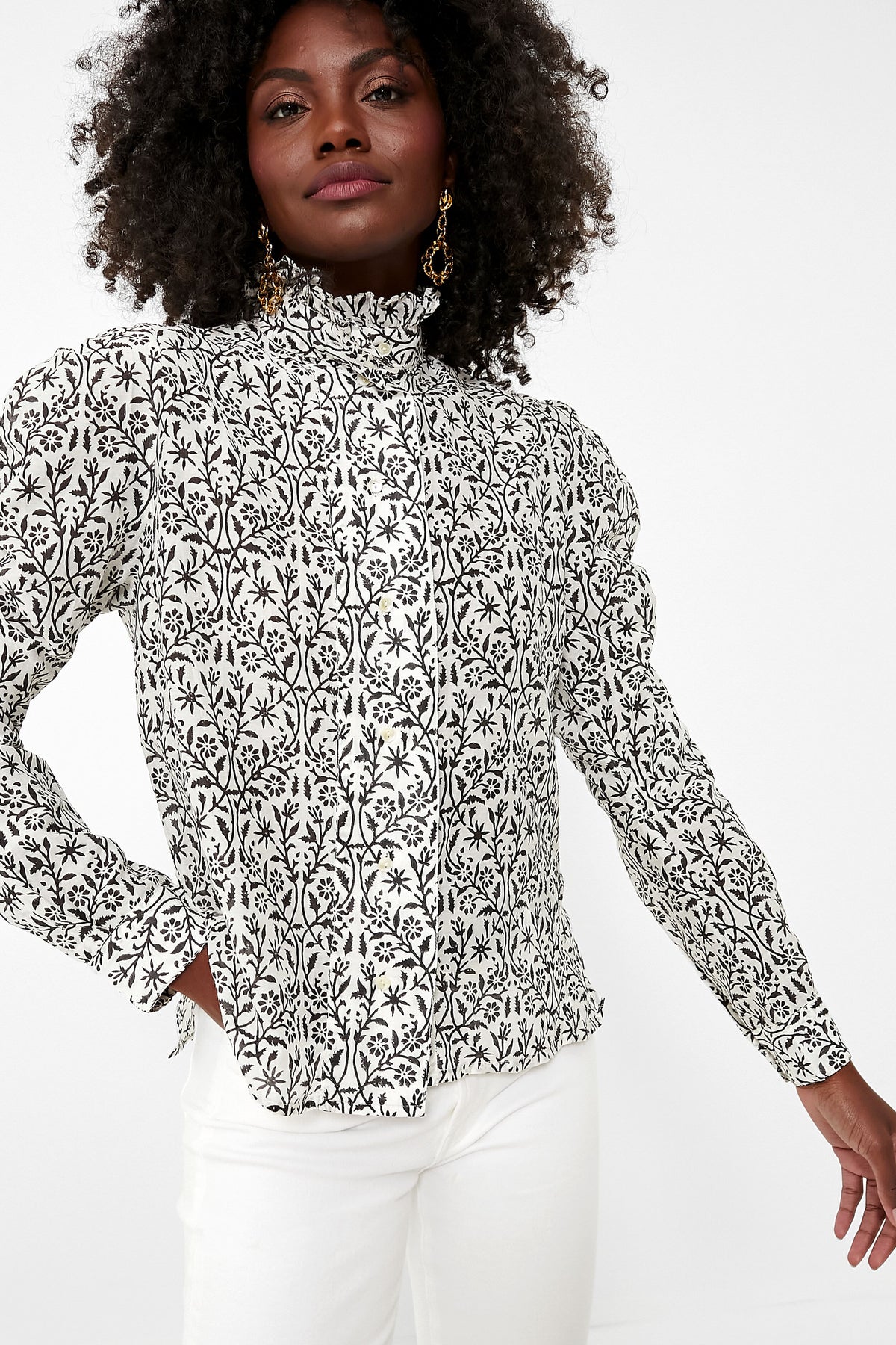 A woman wears a Thierry Colson high-neck, long-sleeve cotton poplin blouse with a black and white floral pattern, paired with white pants.