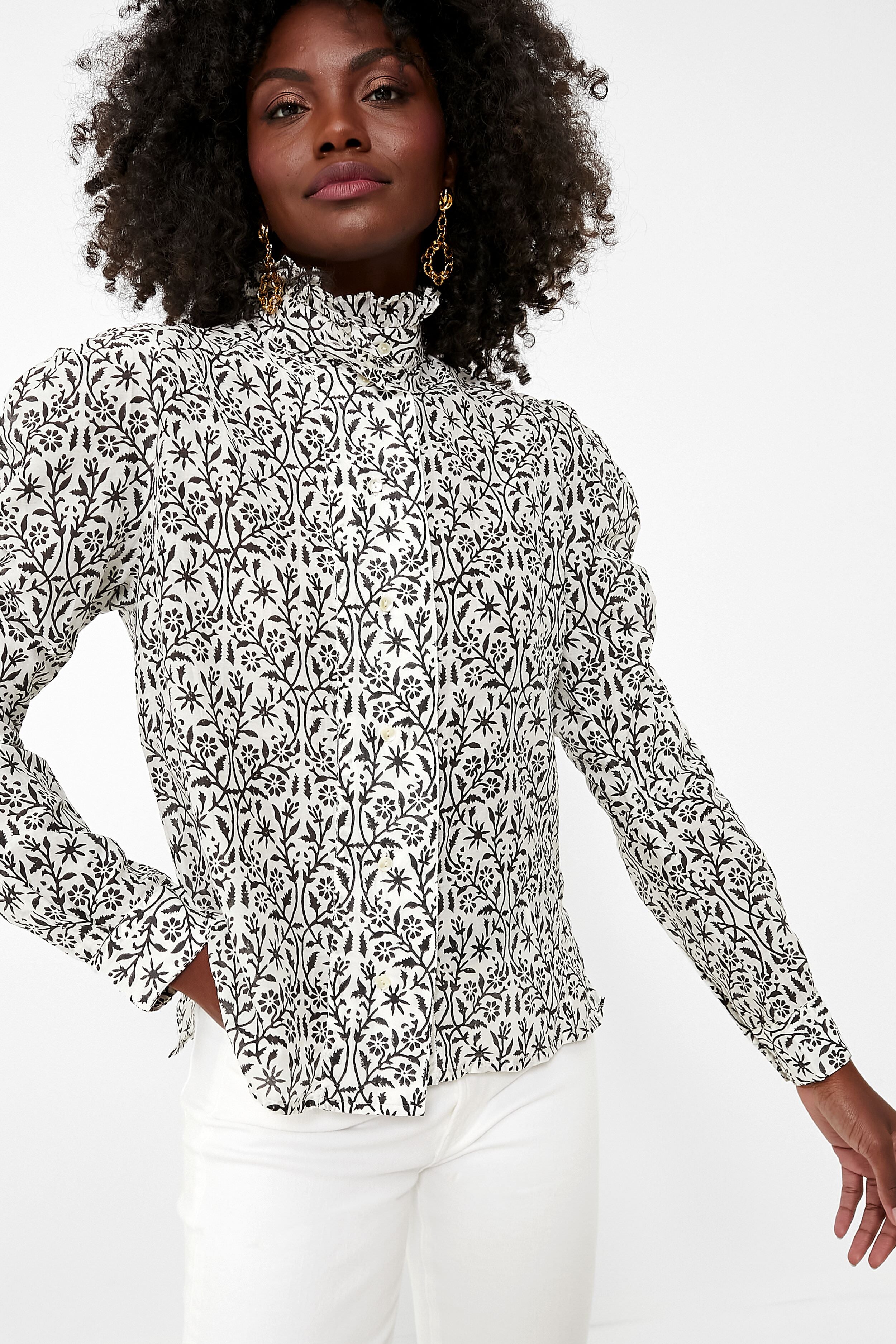A woman wears a Thierry Colson high-neck, long-sleeve cotton poplin blouse with a black and white floral pattern, paired with white pants.