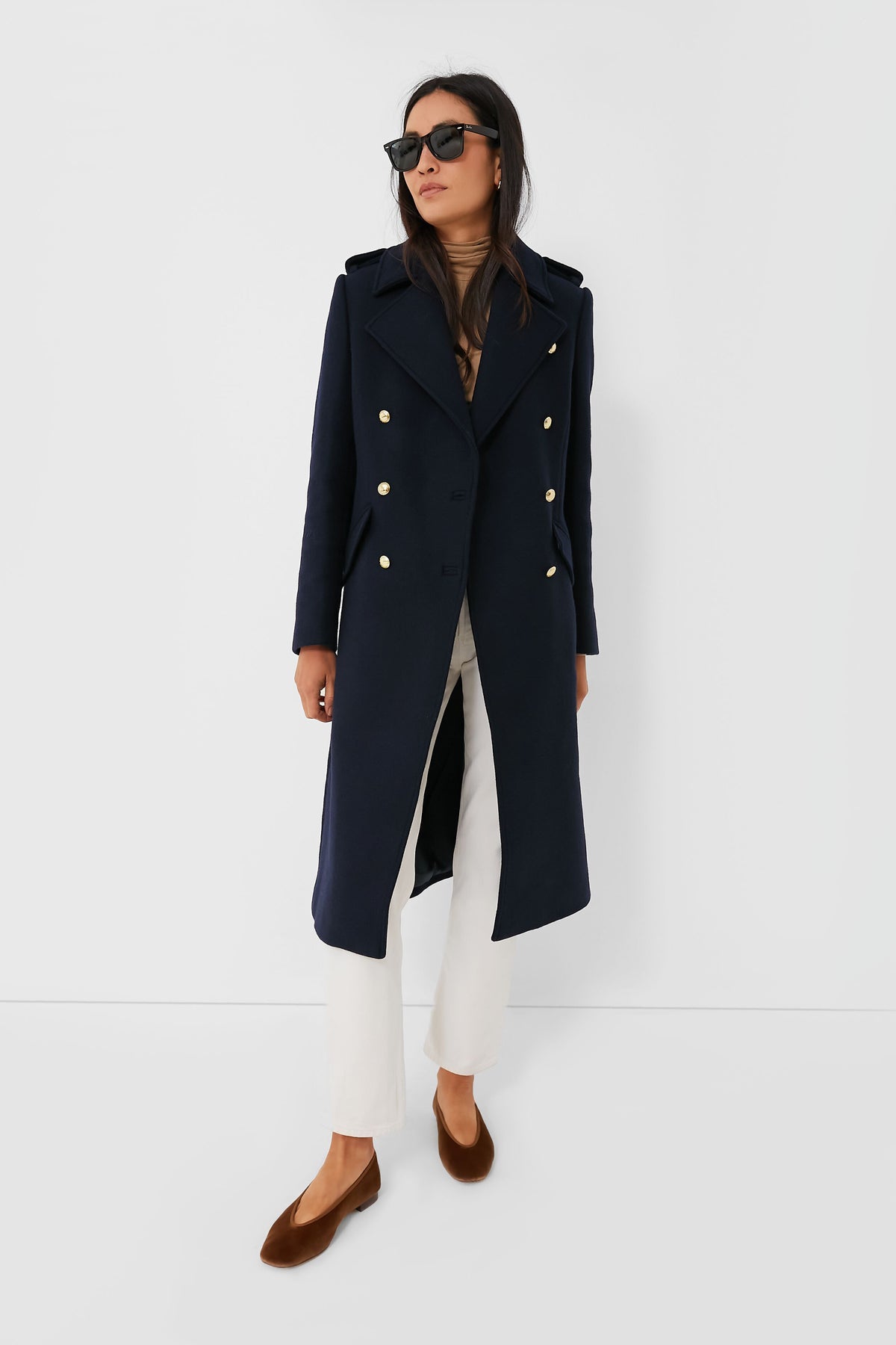 A woman stands confidently wearing Barbour sunglasses, a navy wool coat with gold buttons, a beige turtleneck, white pants, and brown flats.