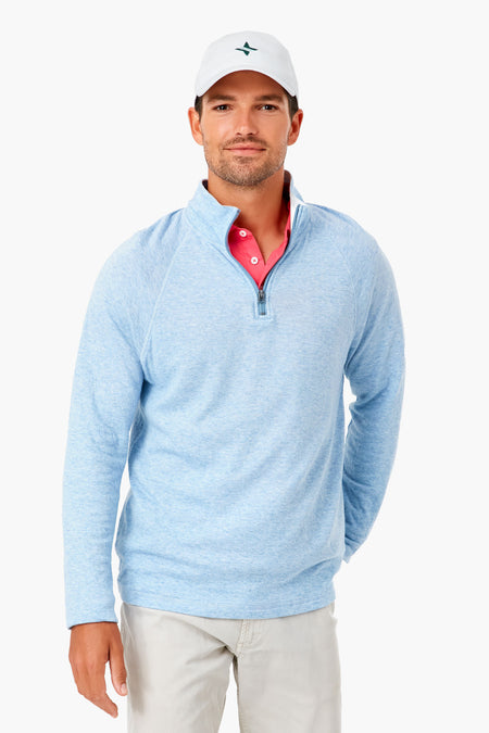 A man in a white cap, Faherty light blue men's casual pullover, pink collared shirt, and beige pants displays a stylish look. The pima cotton quarter zip by Faherty provides comfort and eco-friendly appeal to his outfit.
