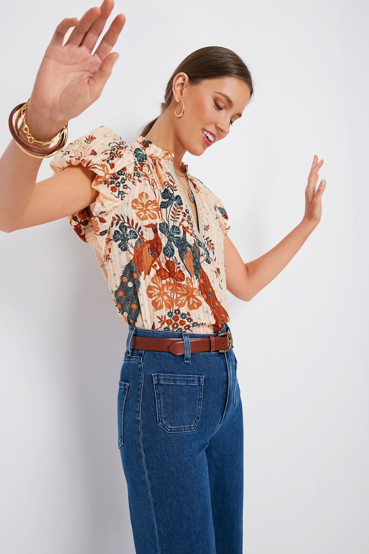A woman smiles with her arms raised and eyes closed, wearing a Ulla Johnson feminine blouse and high-waisted blue jeans.