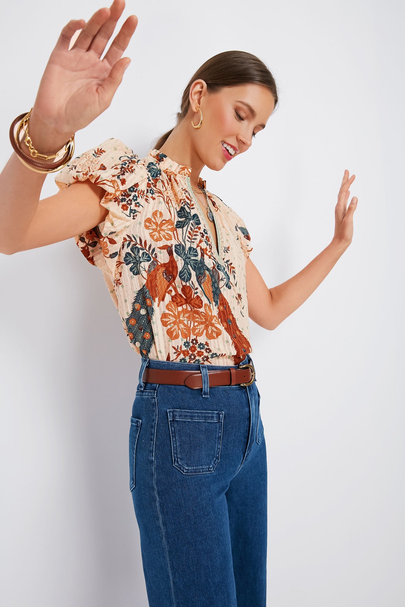 A woman smiles with her arms raised and eyes closed, wearing a Ulla Johnson feminine blouse and high-waisted blue jeans.