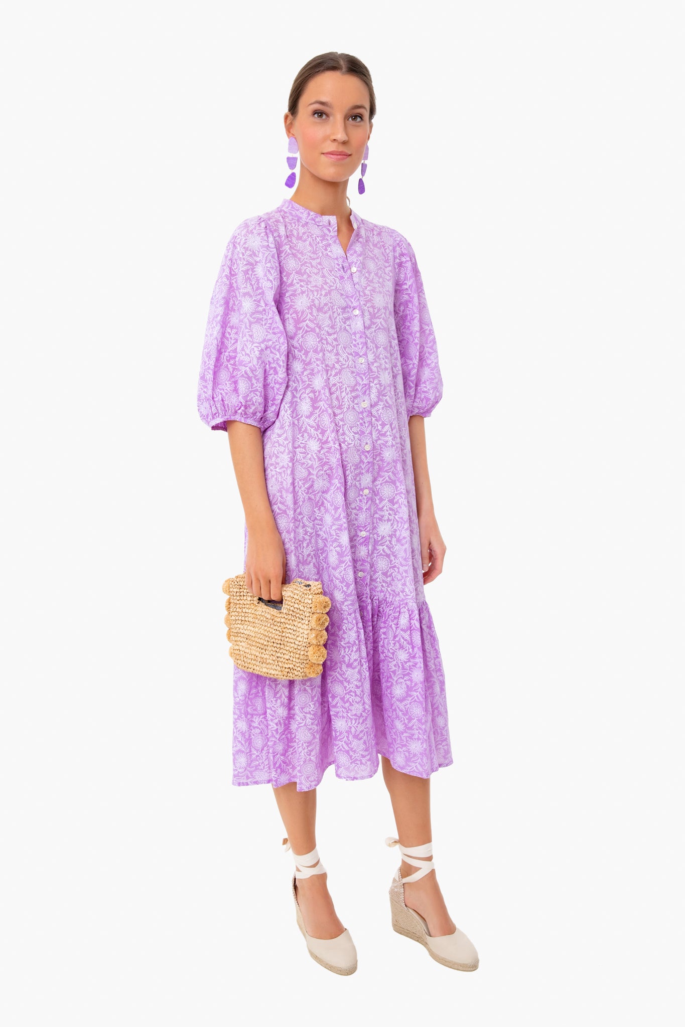 A woman in a loose-fitting, light purple floral midi dress by Ro's Garden with botanical block print, white espadrille wedges, purple earrings, and a woven clutch stands confidently.