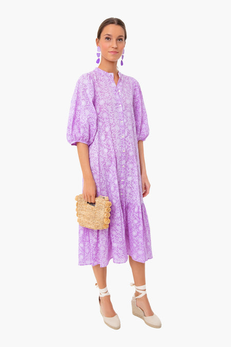 A woman in a loose-fitting, light purple floral midi dress by Ro's Garden with botanical block print, white espadrille wedges, purple earrings, and a woven clutch stands confidently.
