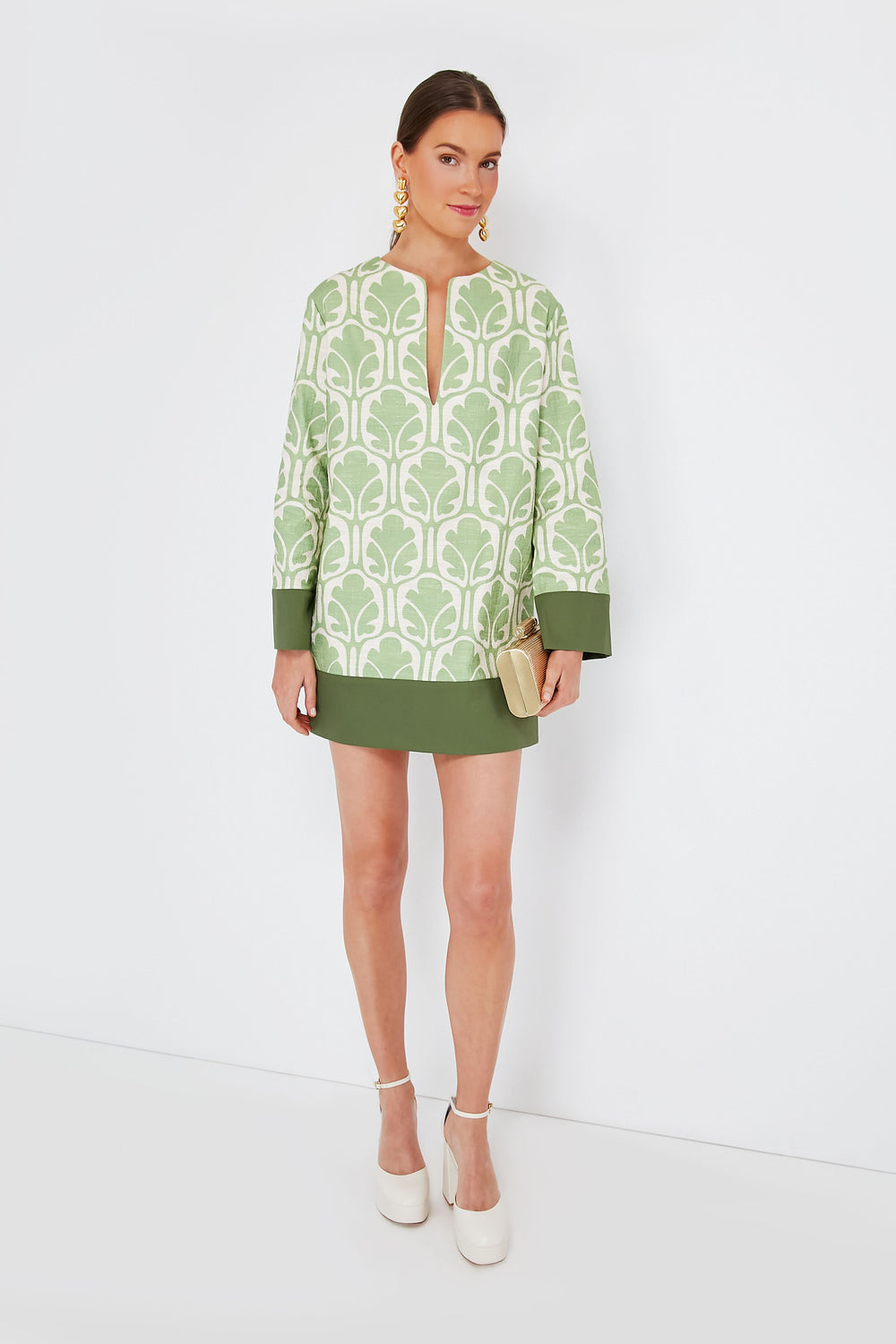 A woman wears La DoubleJ's Grove Deck Mini Dress—a green and white patterned, linen-feel cotton dress—paired with white platform shoes, gold earrings, and a beige clutch. This Italian-made look blends chic style with effortless elegance.