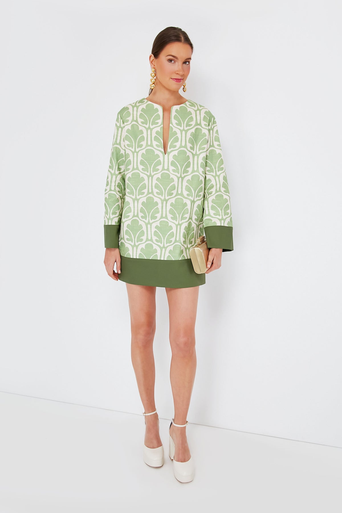 A woman wears La DoubleJ's Grove Deck Mini Dress—a green and white patterned, linen-feel cotton dress—paired with white platform shoes, gold earrings, and a beige clutch. This Italian-made look blends chic style with effortless elegance.