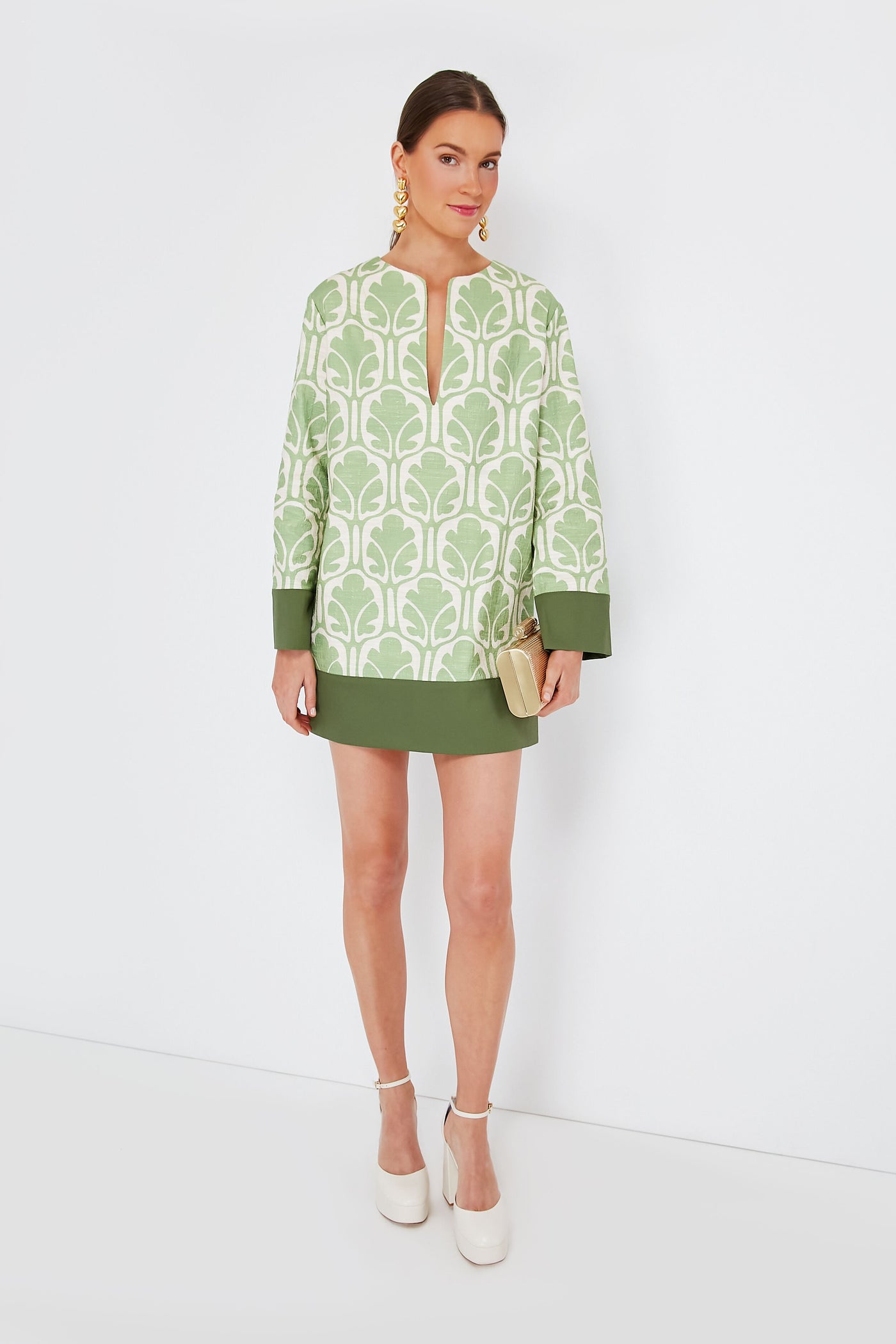 A woman wears La DoubleJ's Grove Deck Mini Dress—a green and white patterned, linen-feel cotton dress—paired with white platform shoes, gold earrings, and a beige clutch. This Italian-made look blends chic style with effortless elegance.