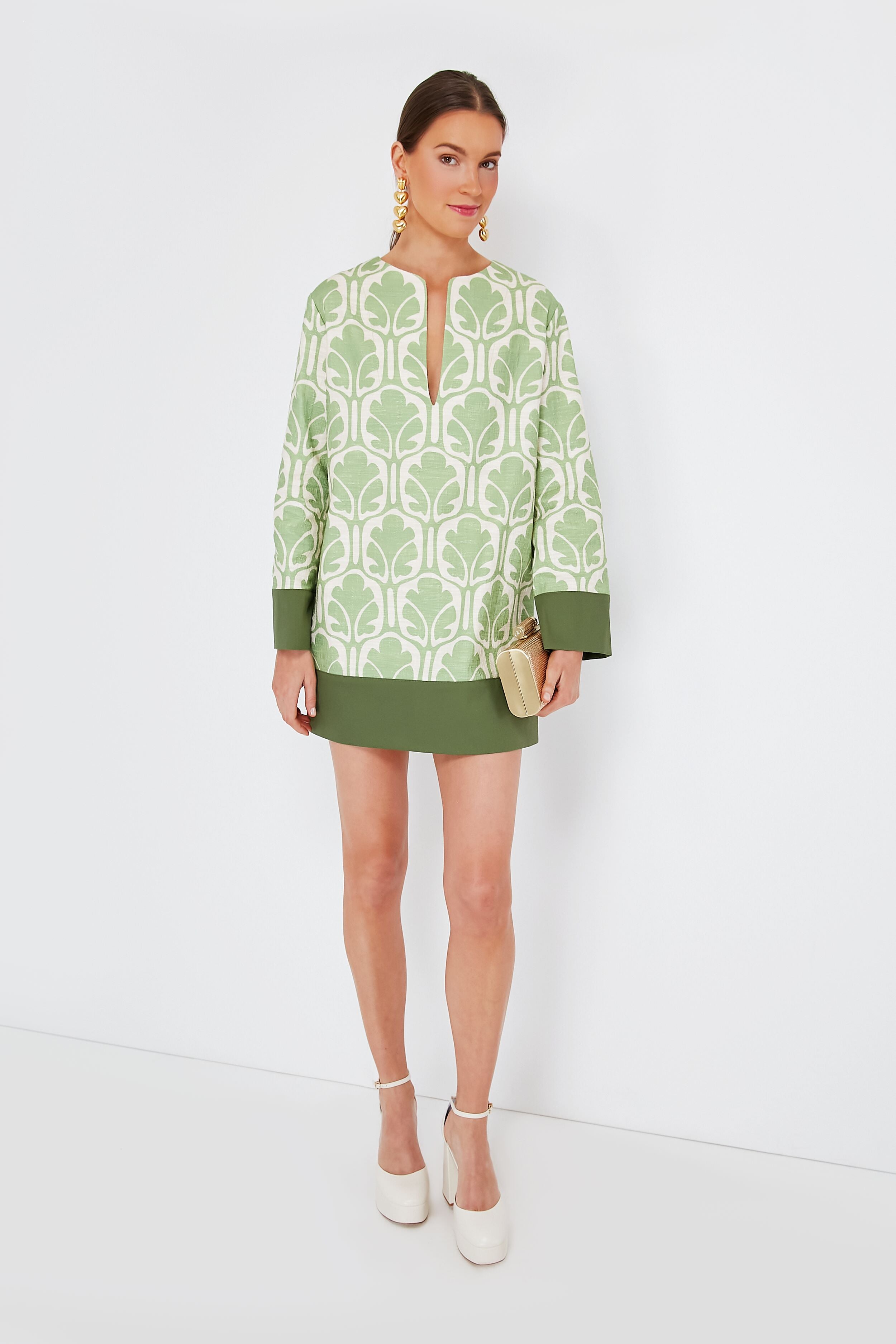 A woman wears La DoubleJ's Grove Deck Mini Dress—a green and white patterned, linen-feel cotton dress—paired with white platform shoes, gold earrings, and a beige clutch. This Italian-made look blends chic style with effortless elegance.