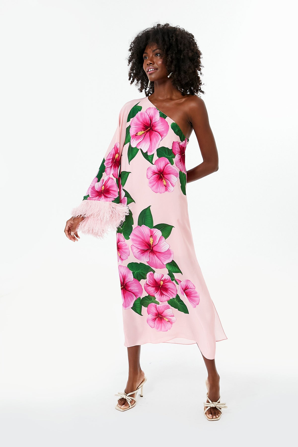 A woman wears a Borgo de Nor Pink Aubrey Dress with an asymmetrical neckline, large pink flower prints, and feather trim on one sleeve, paired with white sandals.