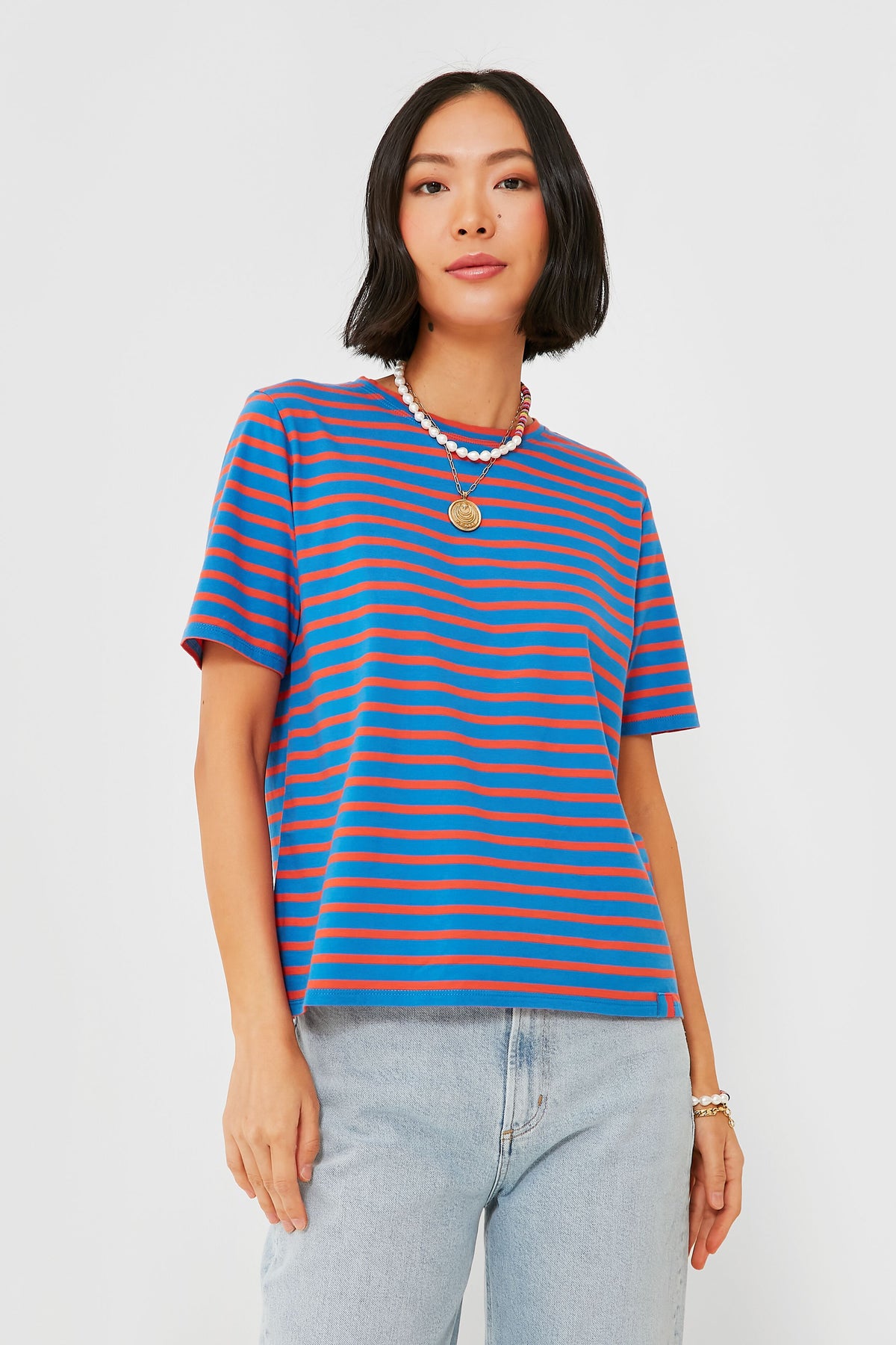 A woman stands confidently in a KULE blue and red stripe tee, light blue jeans, layered necklaces, and bracelets.