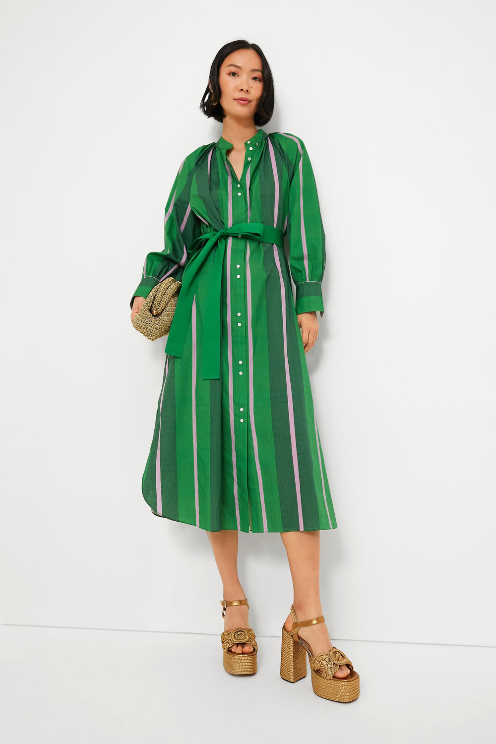 A woman wears the Oroton Jewel Green Calypso Shirt Dress, a striped cotton poplin dress with a tie belt, accessorized with a woven clutch and gold platform sandals.