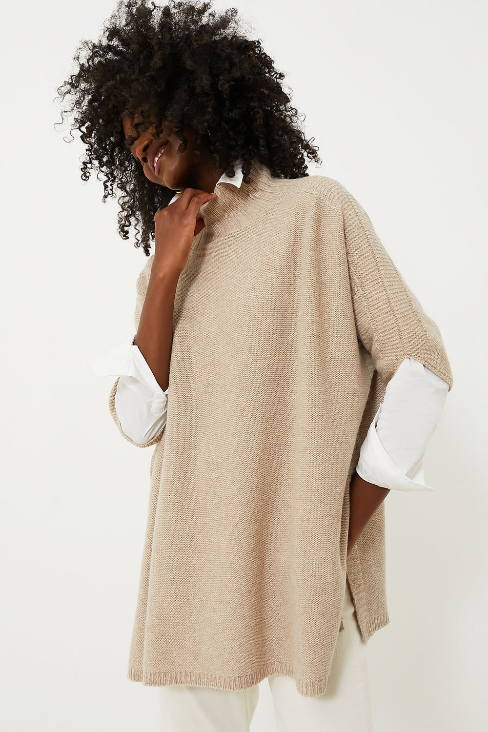 A person in a beige oversized Marella knit sweater, inspired by Italian fashion, layered over a white collared shirt and posing with one hand near their chin.