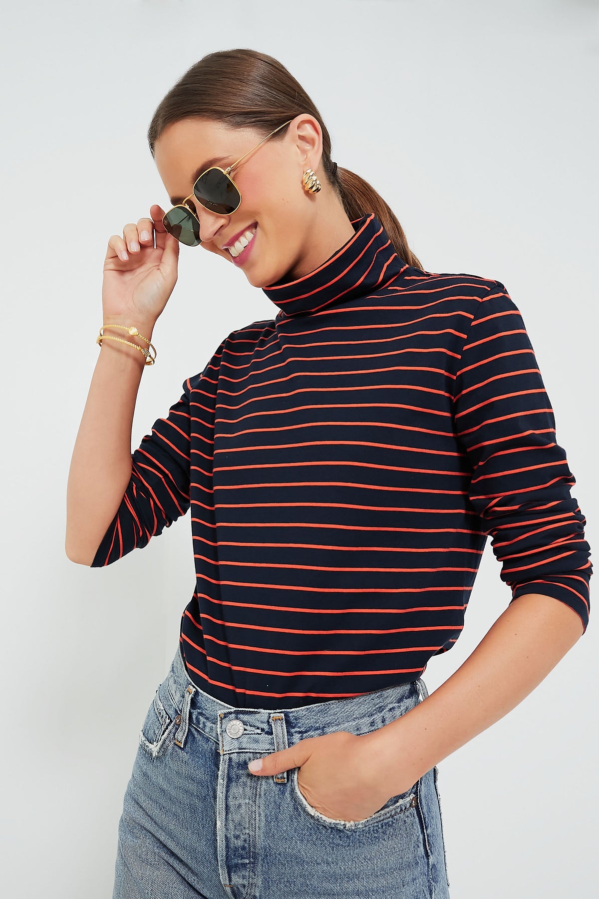 A person in a KULE navy and red striped turtleneck and jeans smiles while touching their sunglasses.