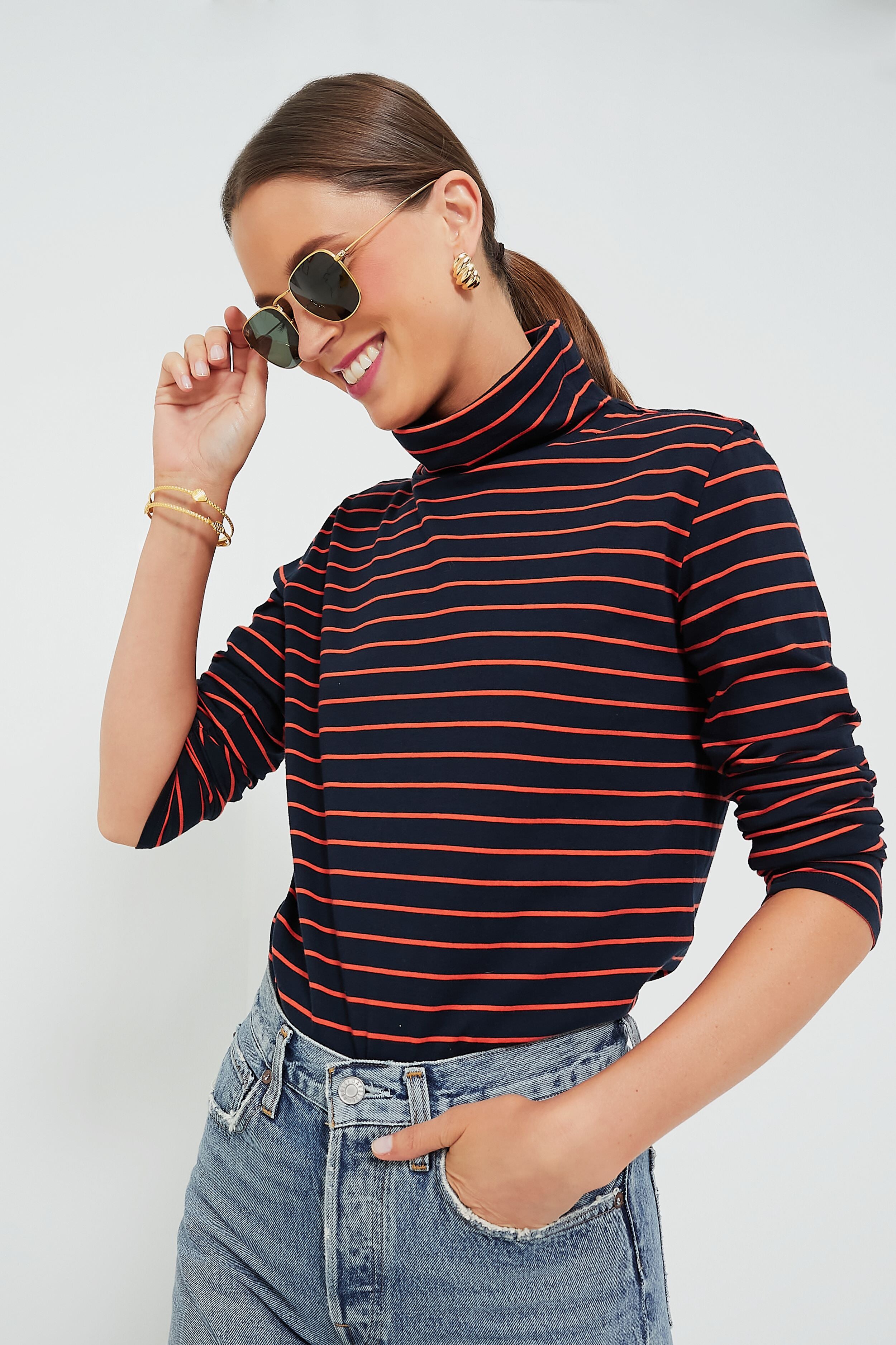 A person in a KULE navy and red striped turtleneck and jeans smiles while touching their sunglasses.