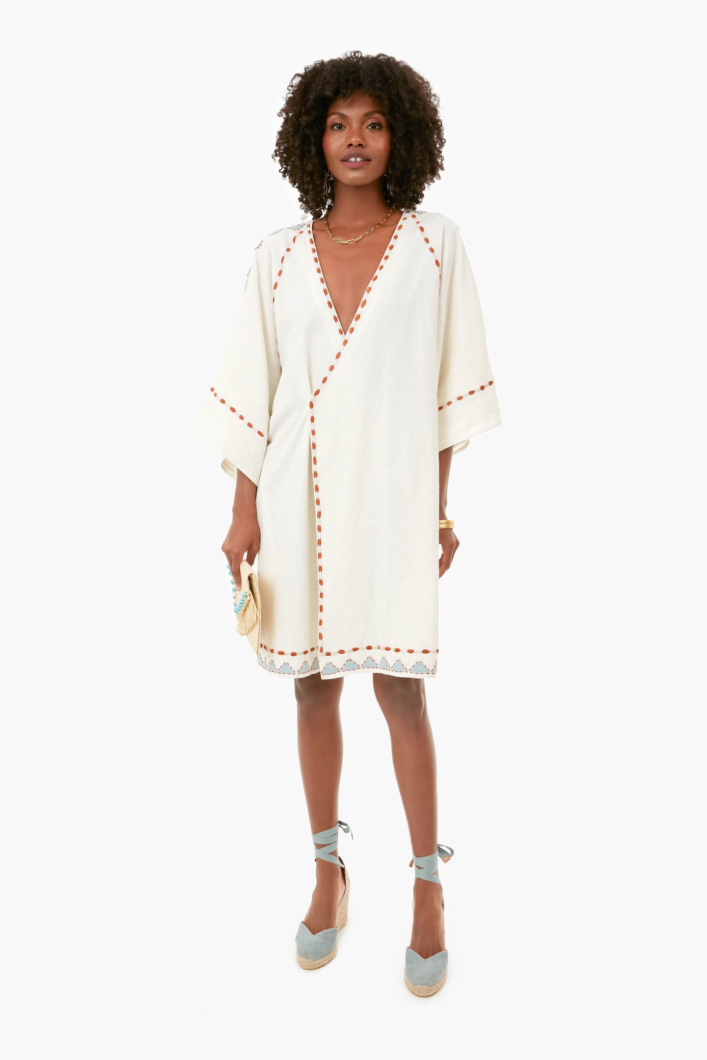 A woman wears a white Carolina K. knee-length vacation dress with wide sleeves, orange trim, lace hem, and blue espadrille wedges, holding a small beige clutch.