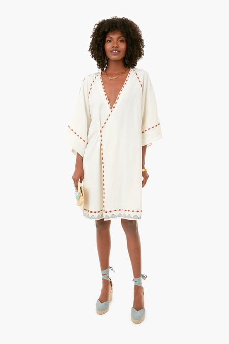 A woman wears a white Carolina K. knee-length vacation dress with wide sleeves, orange trim, lace hem, and blue espadrille wedges, holding a small beige clutch.