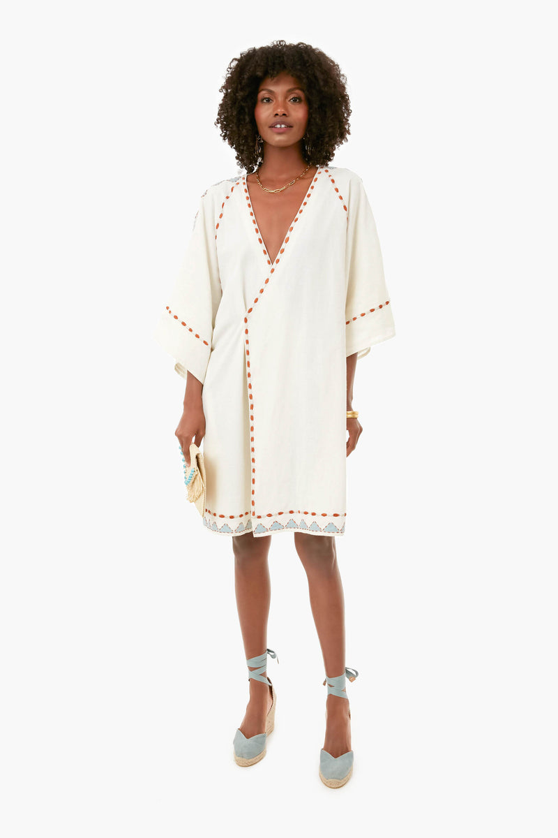 A woman wears a white Carolina K. knee-length vacation dress with wide sleeves, orange trim, lace hem, and blue espadrille wedges, holding a small beige clutch.