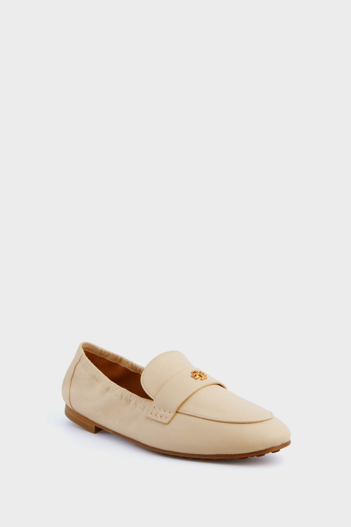 A beige women's leather loafer by Tory Burch, featuring a flat sole and a gold decorative emblem on the upper.