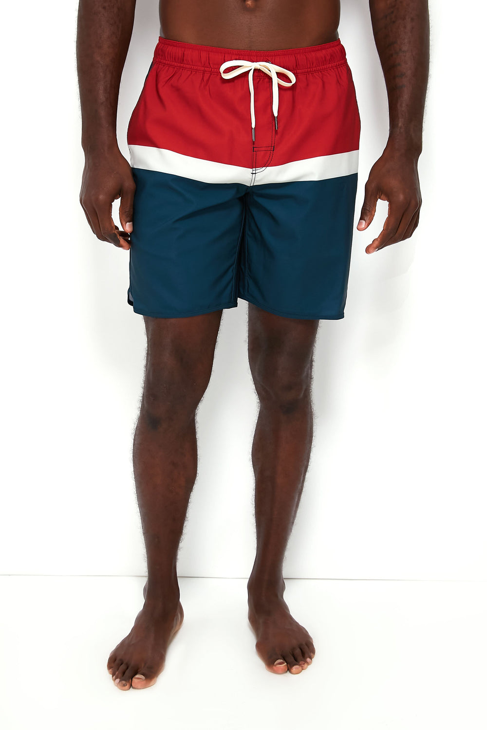 A man stands barefoot wearing Fair Harbor red, white, and blue color-blocked swim trunks made from upcycled plastic bottles, featuring a white drawstring.