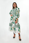 She wears the Sue Sartor Emerald Marigold Flounce Dress, a white midi with green floral patterns, long sleeves, and tied waist. Smiling warmly, her dark hair frames her face as she pairs the look with navy platform shoes.