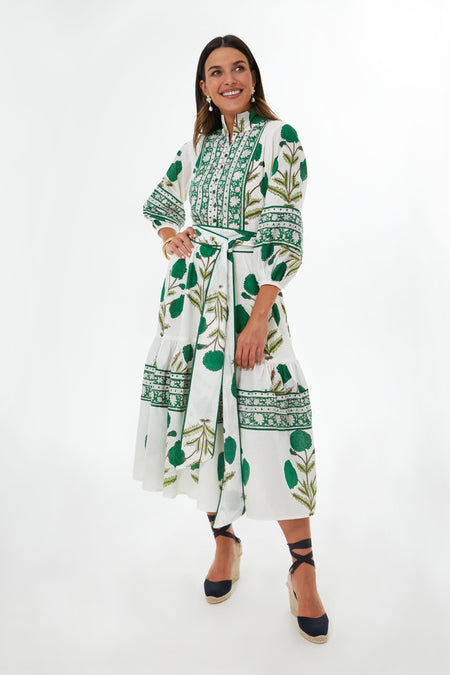 She wears the Sue Sartor Emerald Marigold Flounce Dress, a white midi with green floral patterns, long sleeves, and tied waist. Smiling warmly, her dark hair frames her face as she pairs the look with navy platform shoes.