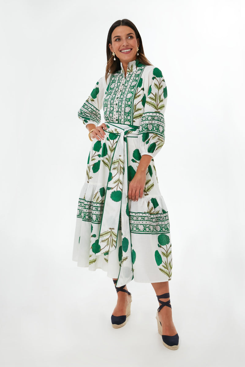 She wears the Sue Sartor Emerald Marigold Flounce Dress, a white midi with green floral patterns, long sleeves, and tied waist. Smiling warmly, her dark hair frames her face as she pairs the look with navy platform shoes.