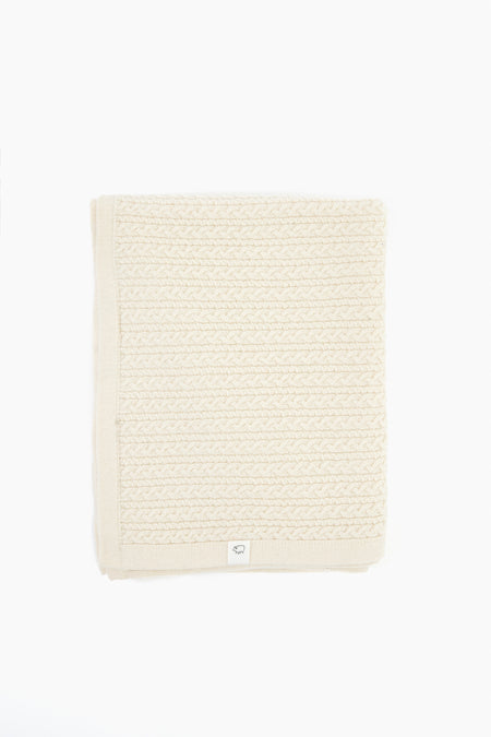 A cream-colored, folded knitted heirloom baby blanket by baa, crafted from Hudson Valley Cormo wool with a textured pattern and finished with a small white tag on one edge.