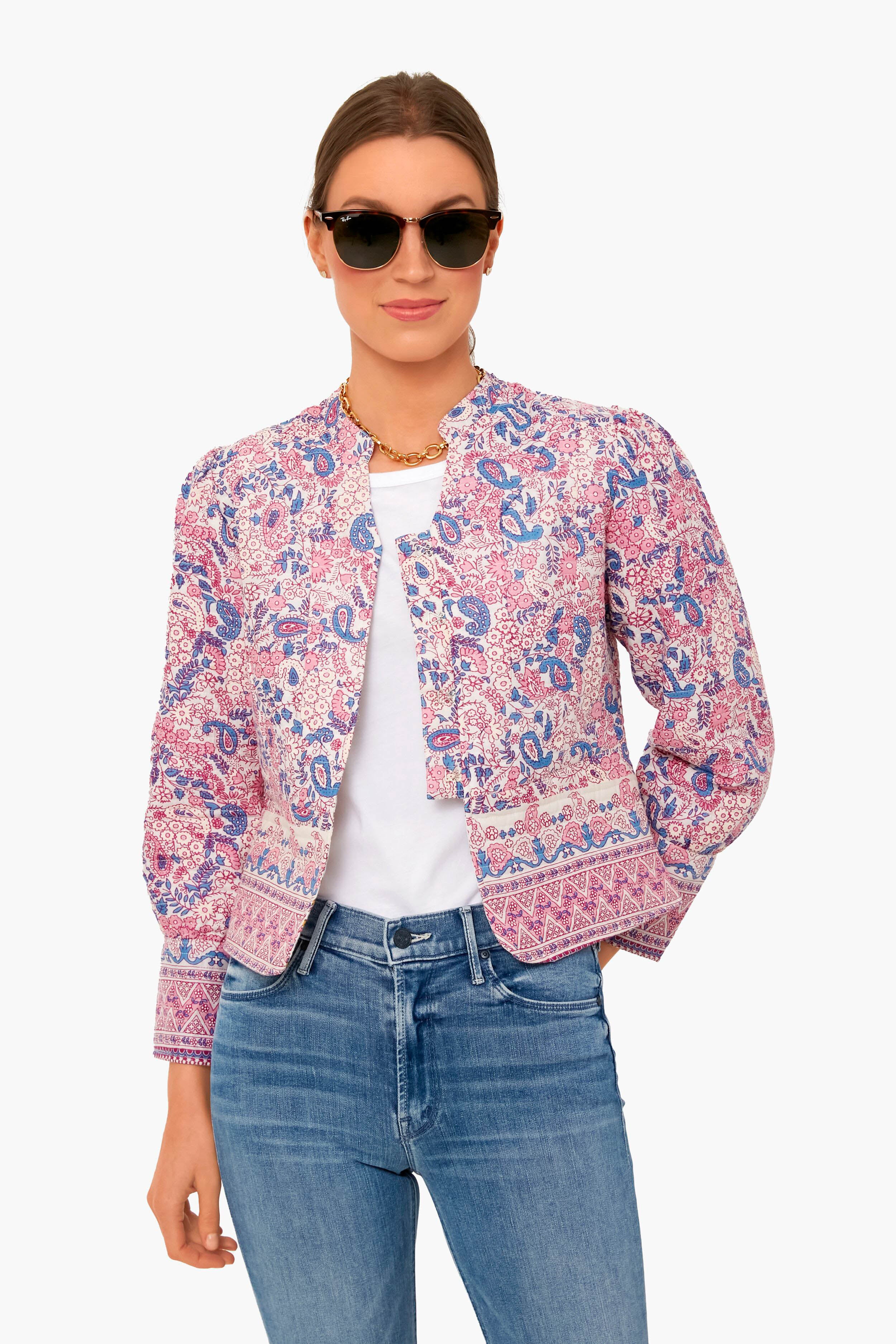 A woman stands confidently wearing Antik Batik sunglasses, a white shirt, a pink and blue batik-printed jacket, and blue jeans.