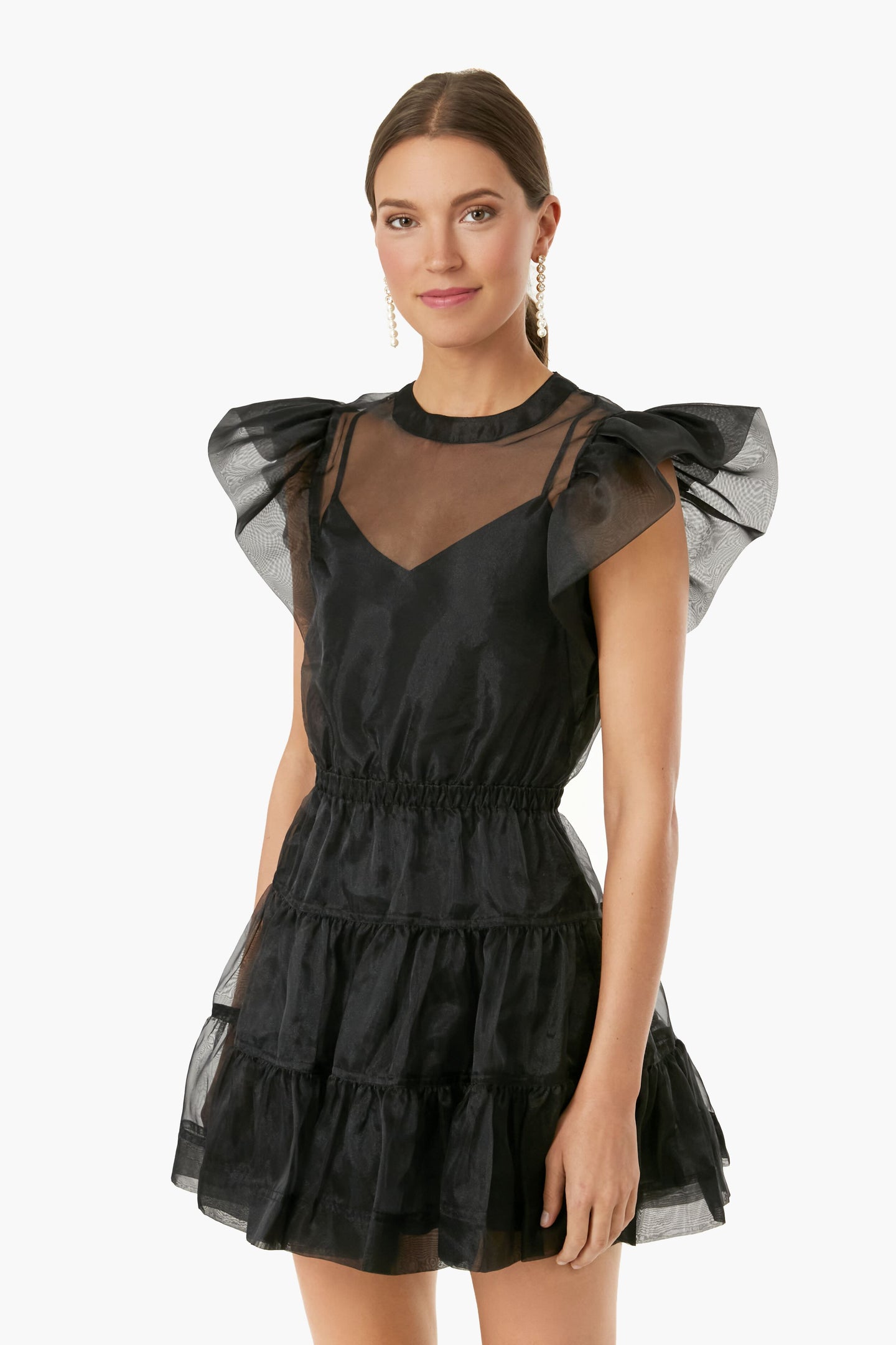 A woman wears a short, black ruffled SIMKHAI organza dress with puff sleeves and a sheer bodice, capturing a luxurious ready-to-wear style.