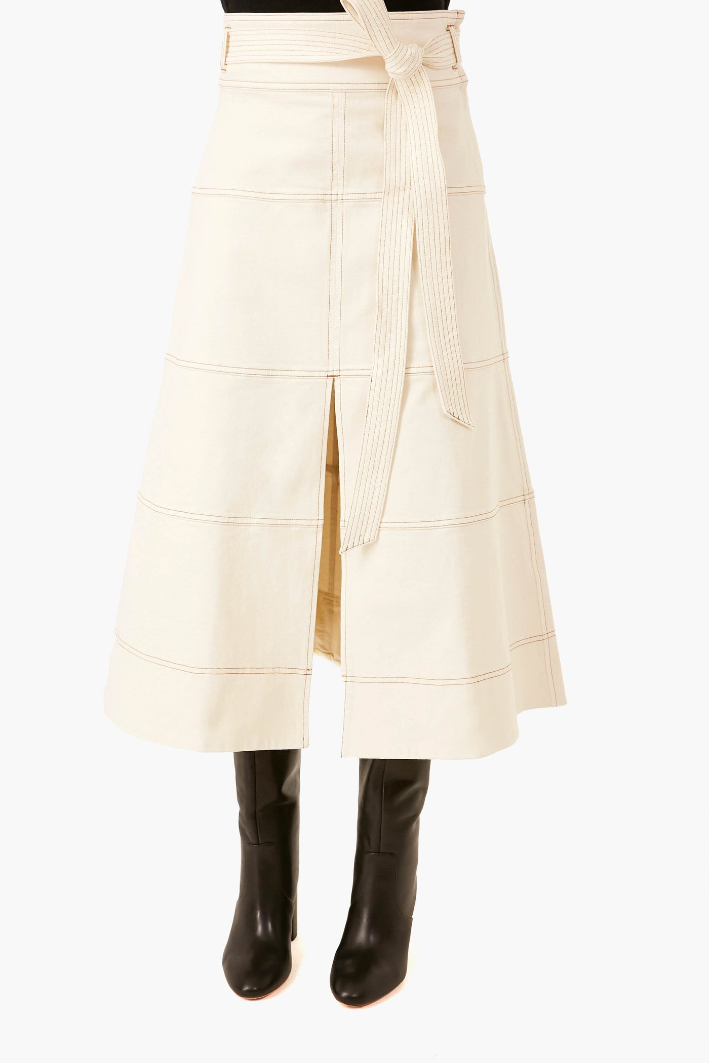 Someone dressed in a Tanya Taylor organic cotton cream belted midi skirt with a front slit, paired with black knee-high boots.
