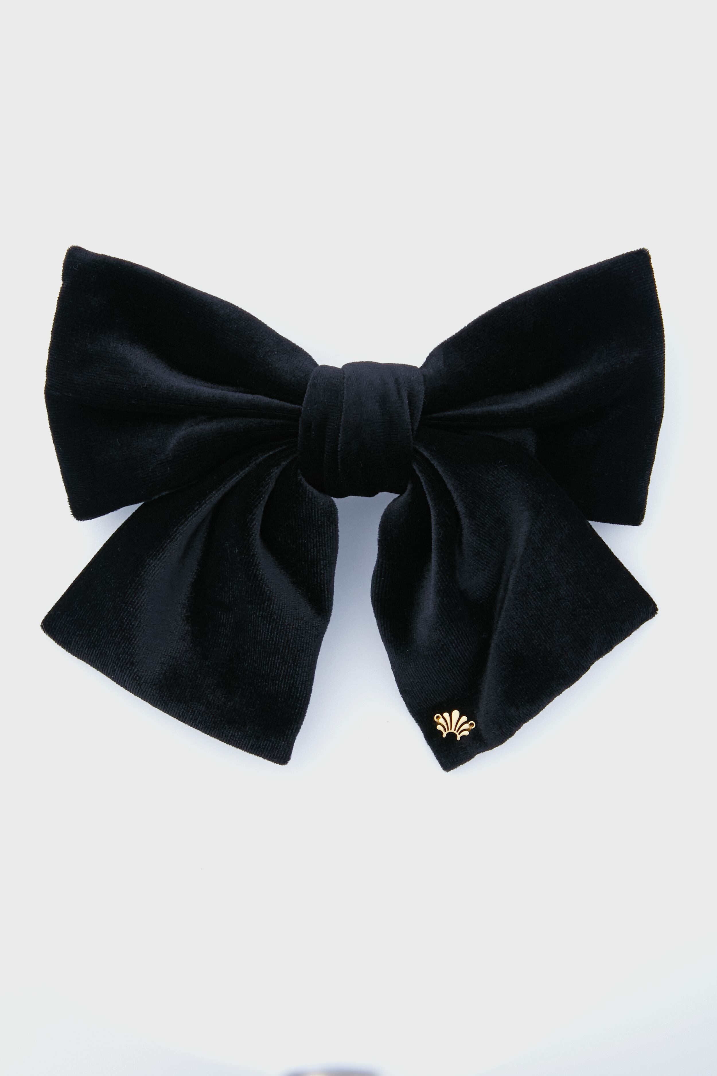 A black velvet hair bow barrette by Lele Sadoughi, featuring a gold emblem on one tail for a stylish touch.