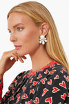 A woman with long blonde hair in profile, wearing a black top with red and white hearts and statement earrings from Tuckernuck Jewelry, rests her hand near her chin.