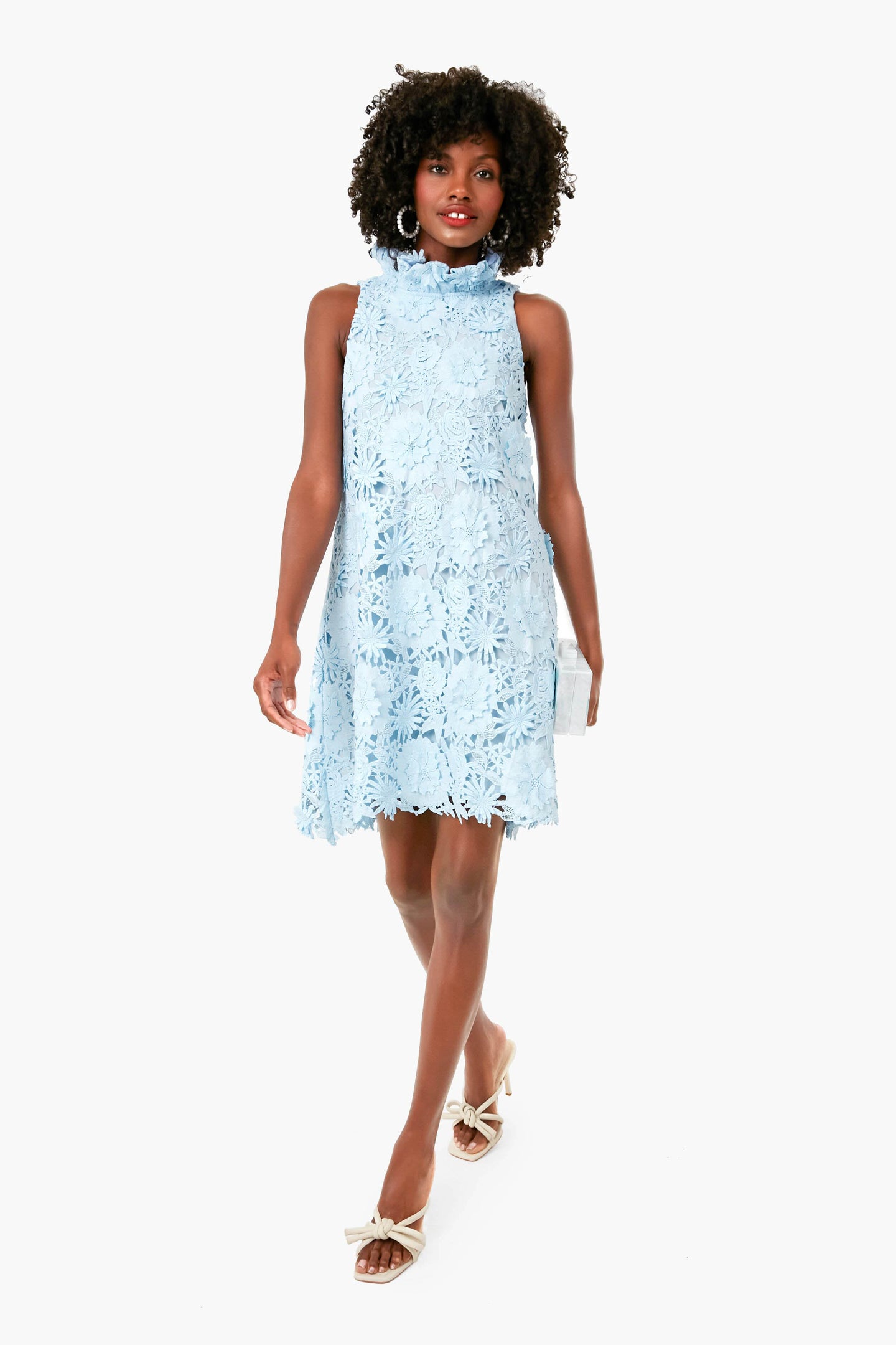 A woman in a Tuckernuck sleeveless light blue lace swing dress with a ruffle stand collar and open-toe bow heels walks forward, carrying a small clutch.