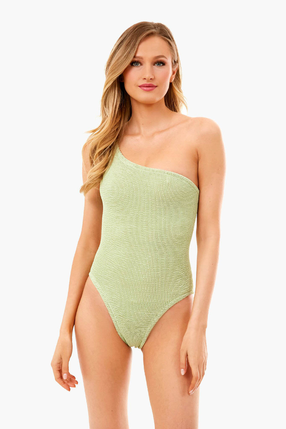 A woman wears a Hunza G light green one-shoulder swimsuit featuring a crinkle-stretch texture and high-cut legs.