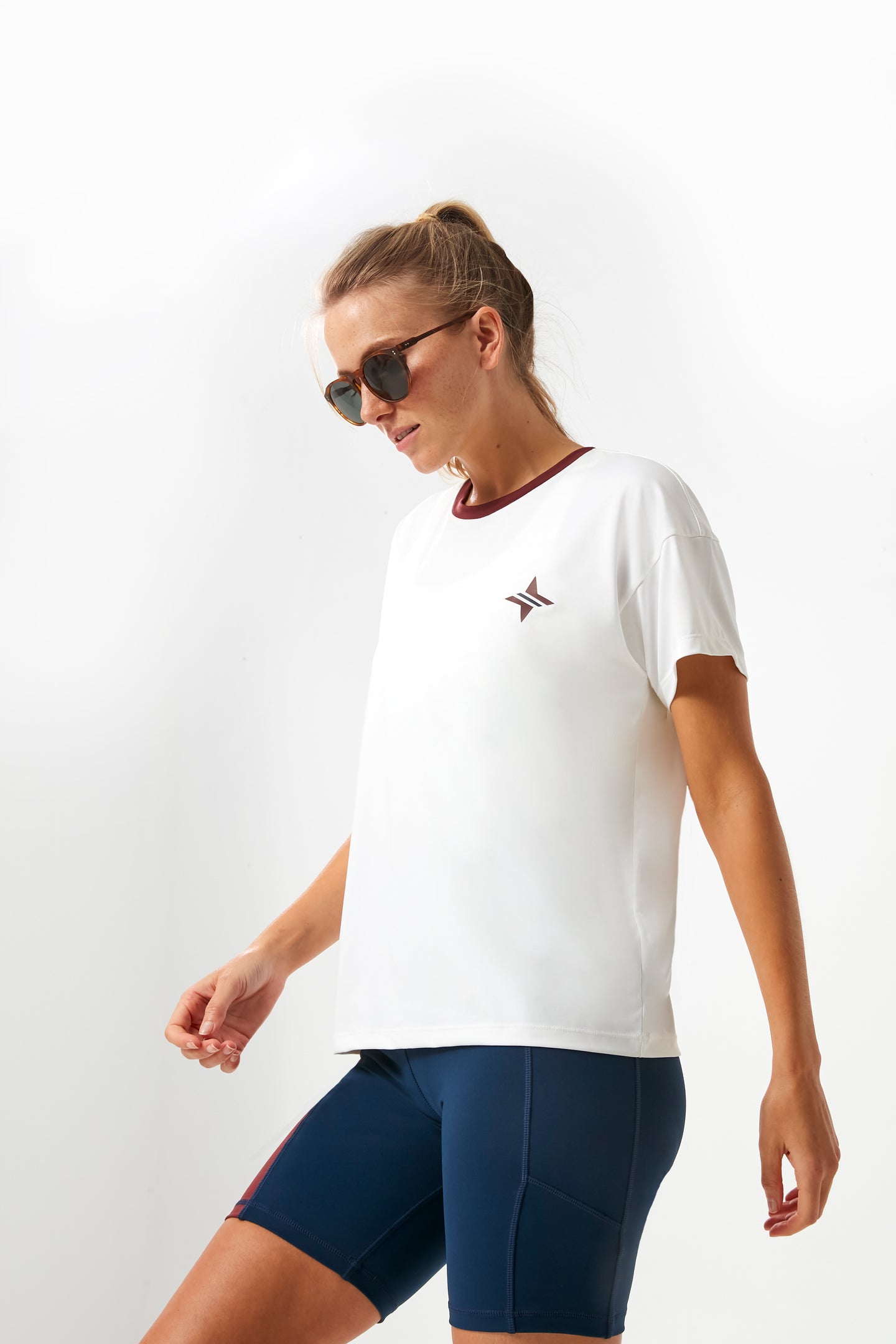 A woman stands confidently in her Tnuck Sport boyfriend tee, wearing sunglasses, a white collegiate t-shirt with a small logo, and navy blue shorts.